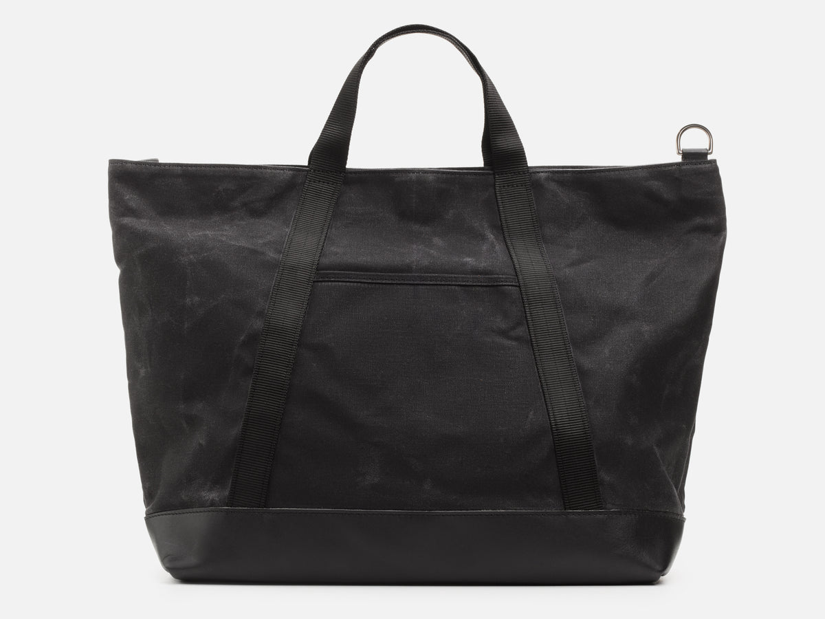 A back view of an black waxed canvas tote bag with 1" nylon black handles with pocket between the handles, and a black leather bottom, set against a plain white background.