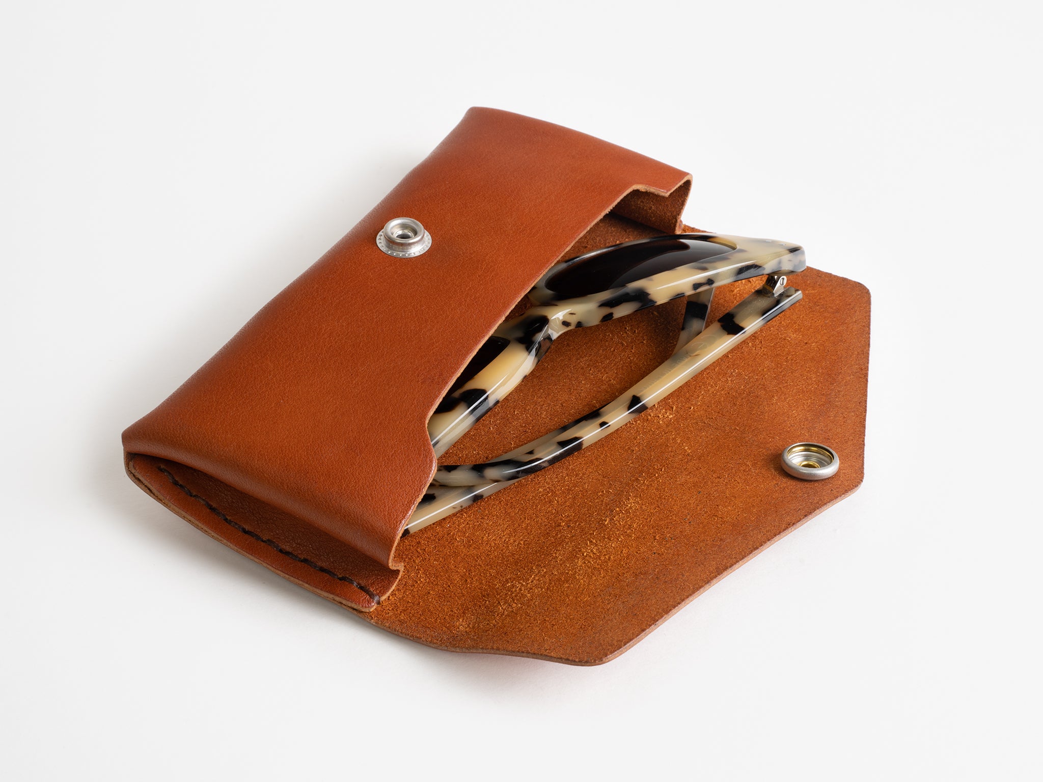 A brown leather case with a snap closure, partially open to reveal a pair of tortoiseshell-patterned eyeglasses inside, all placed on a white background.