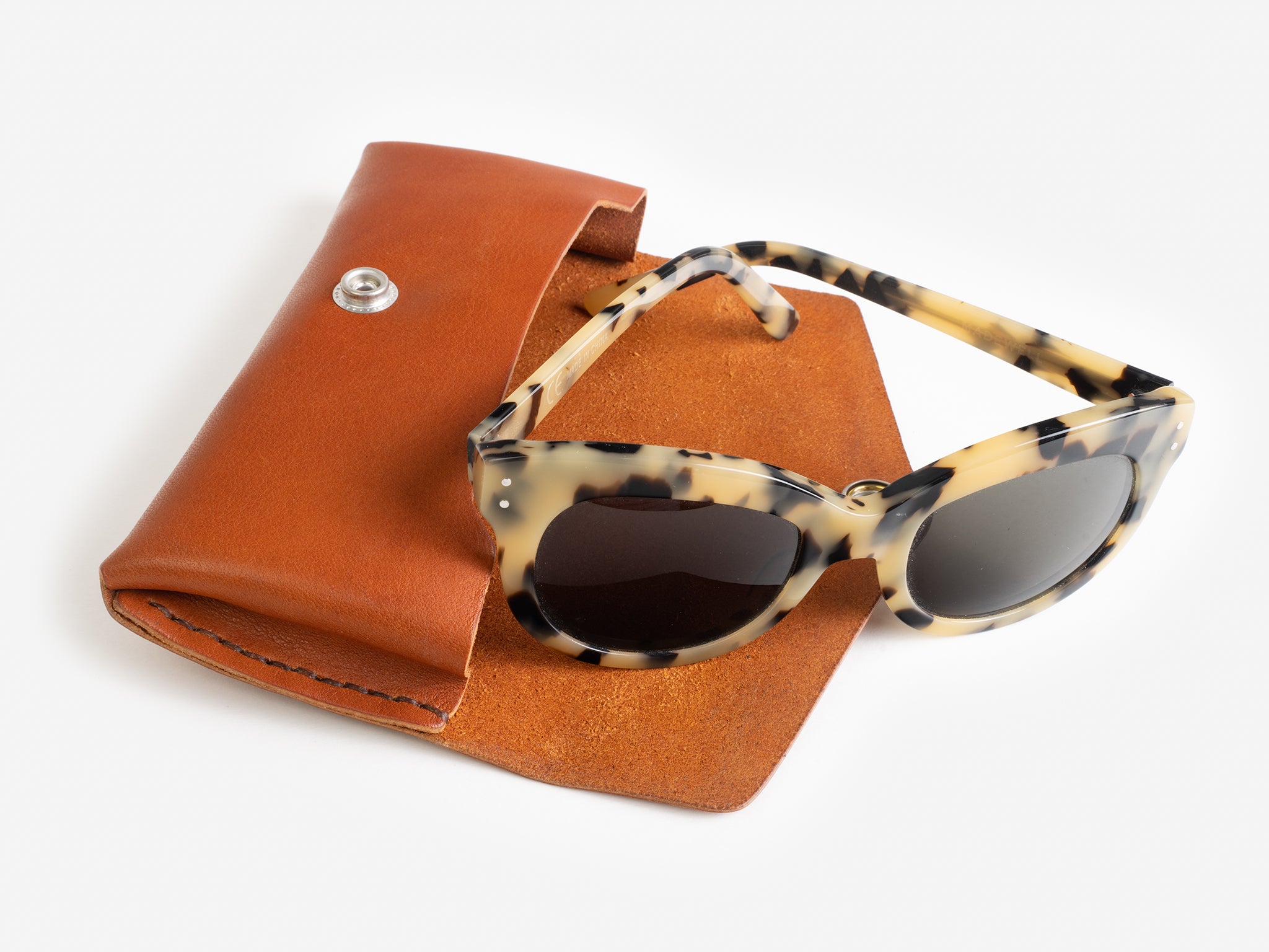 Tortoiseshell sunglasses with dark lenses rest on an open brown leather case with a snap button, displayed on a white background.