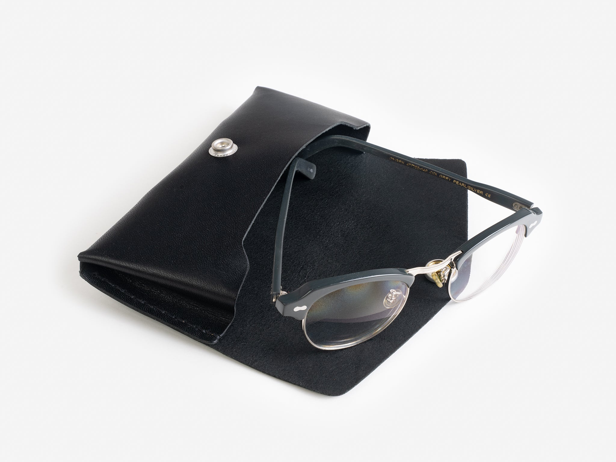 A pair of black-framed eyeglasses with gold accents rests on an open black leather case with a snap closure, set against a white background.