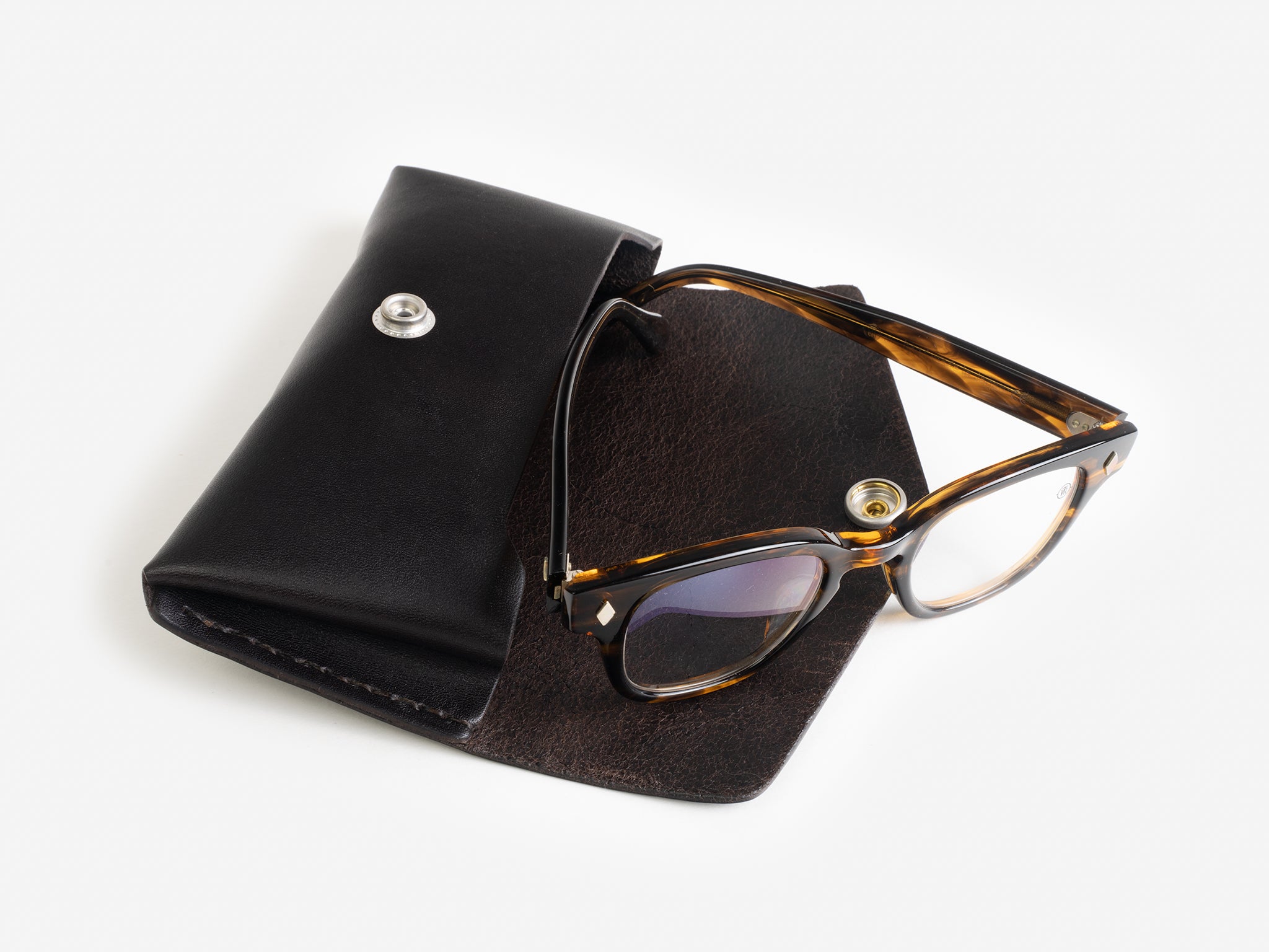 A pair of tortoiseshell eyeglasses partially inside an open black leather case with a snap button, displayed on a white background.