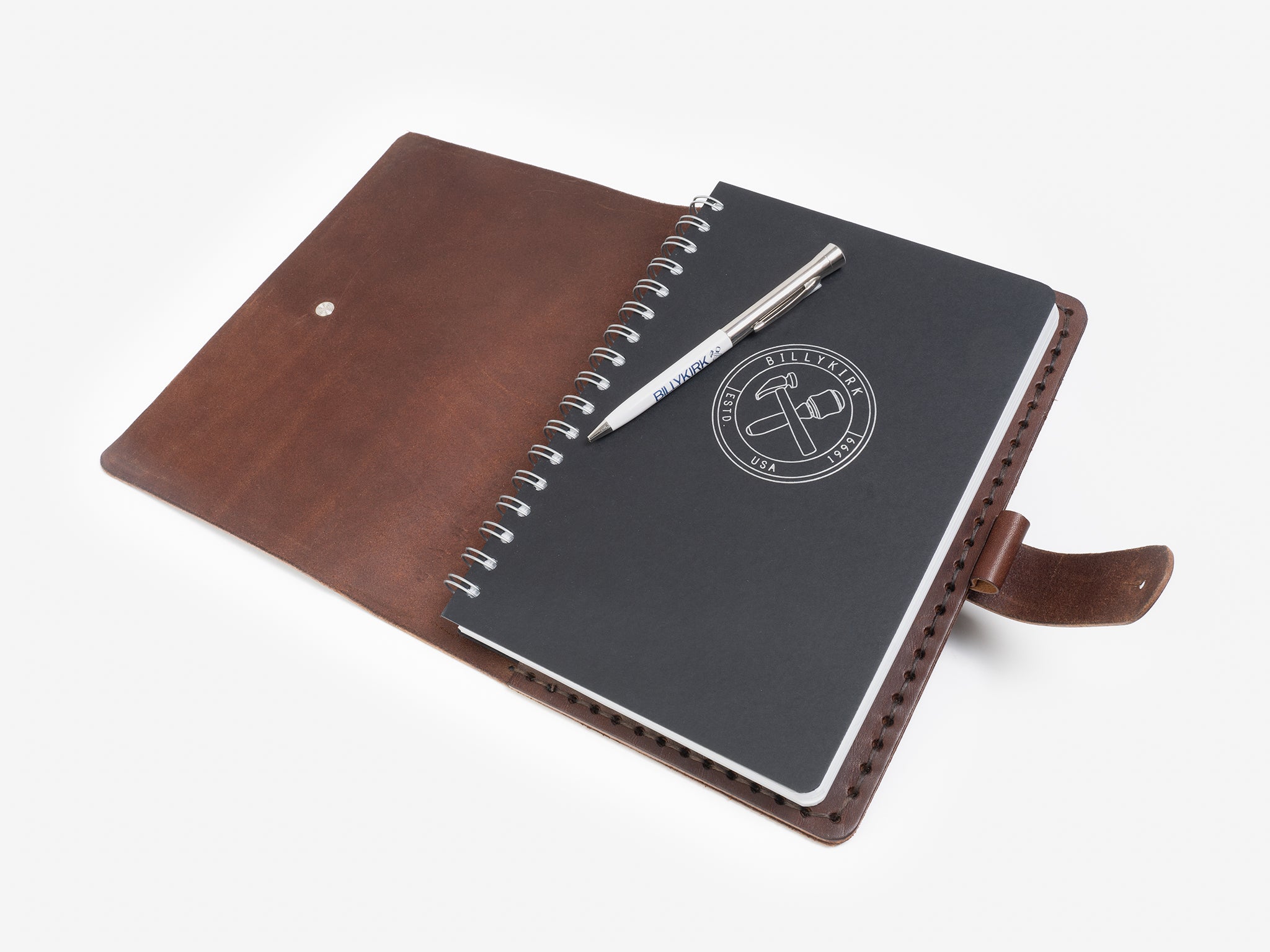 A brown leather folio is open, revealing a black spiral-bound notebook with a logo and a silver pen resting on top, all set against a white background.