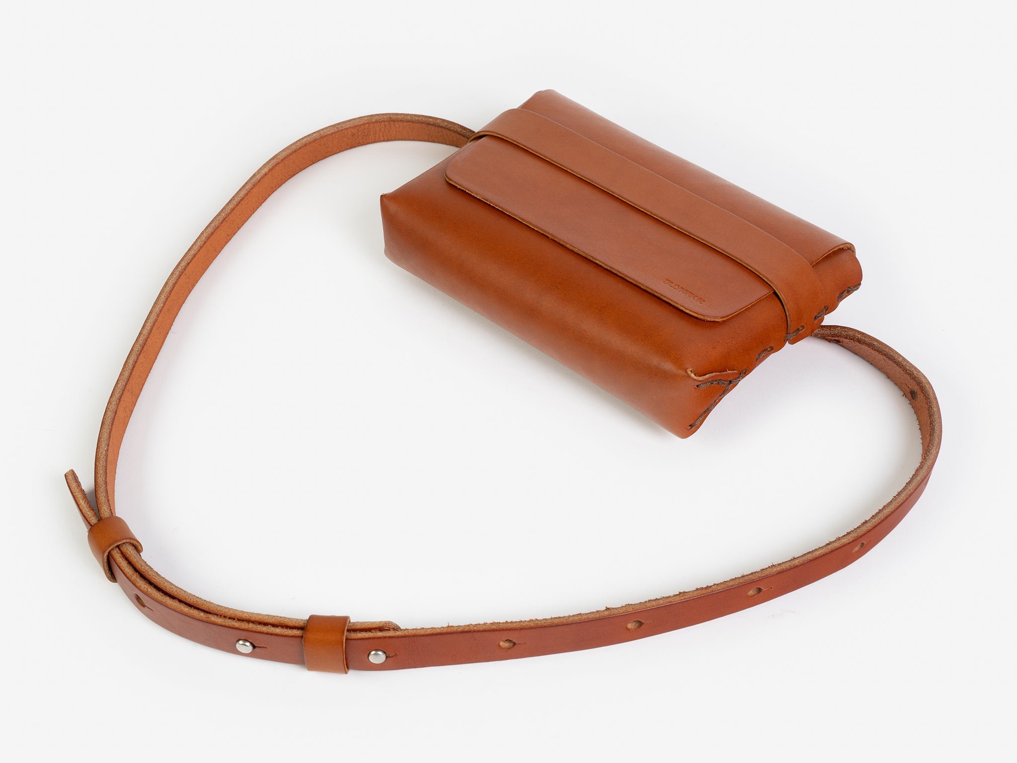 A tan leather belt crossbody bag with a smooth finish, featuring a flap closure, subtle embossed branding, and an adjustable strap with silver-tone collar buttons, displayed against a white background.