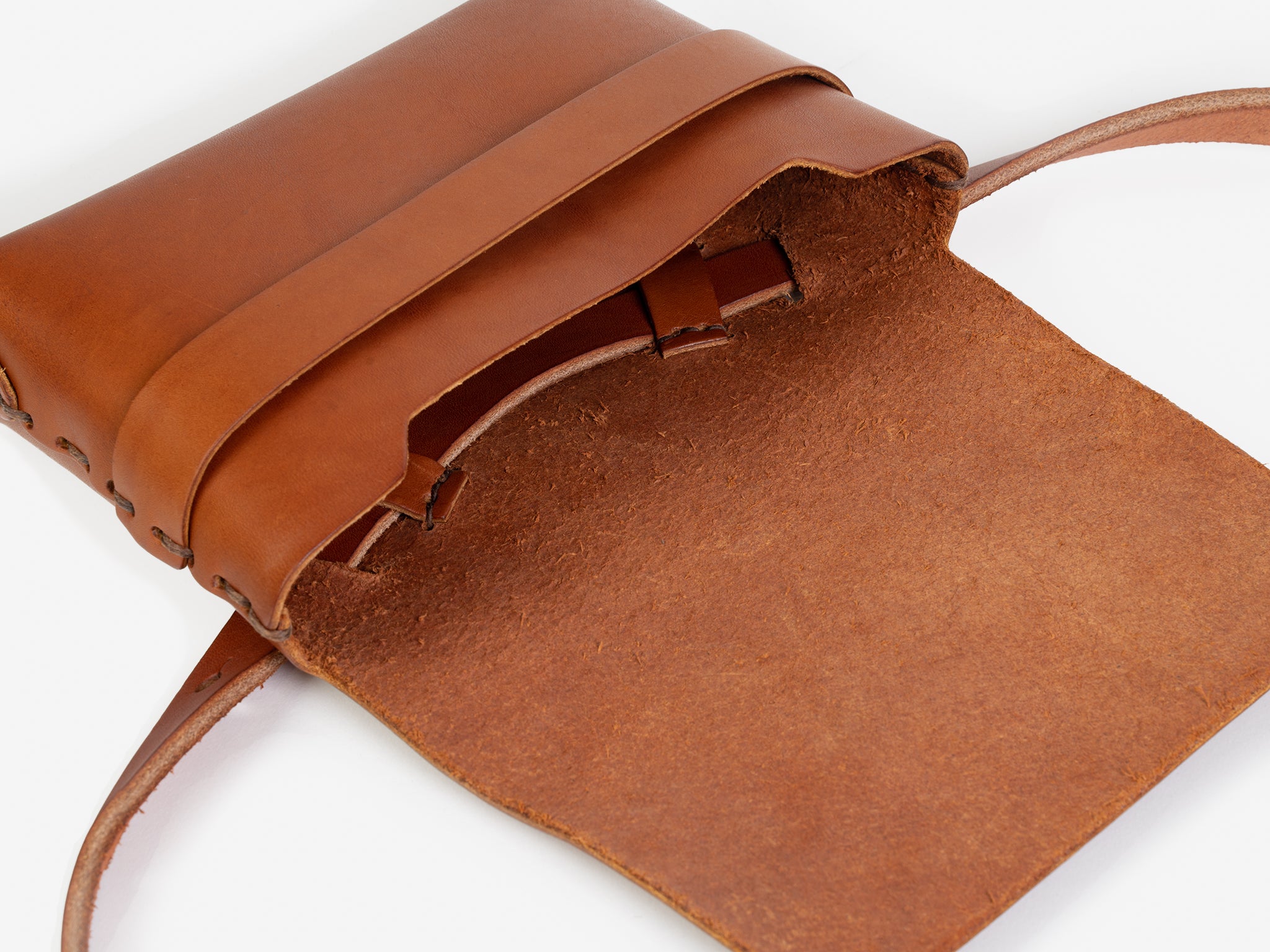 A tan leather crossbody bag with a flap open, revealing some hand-stitched details. The bag has a strap and minimalist design, displayed on a white background.