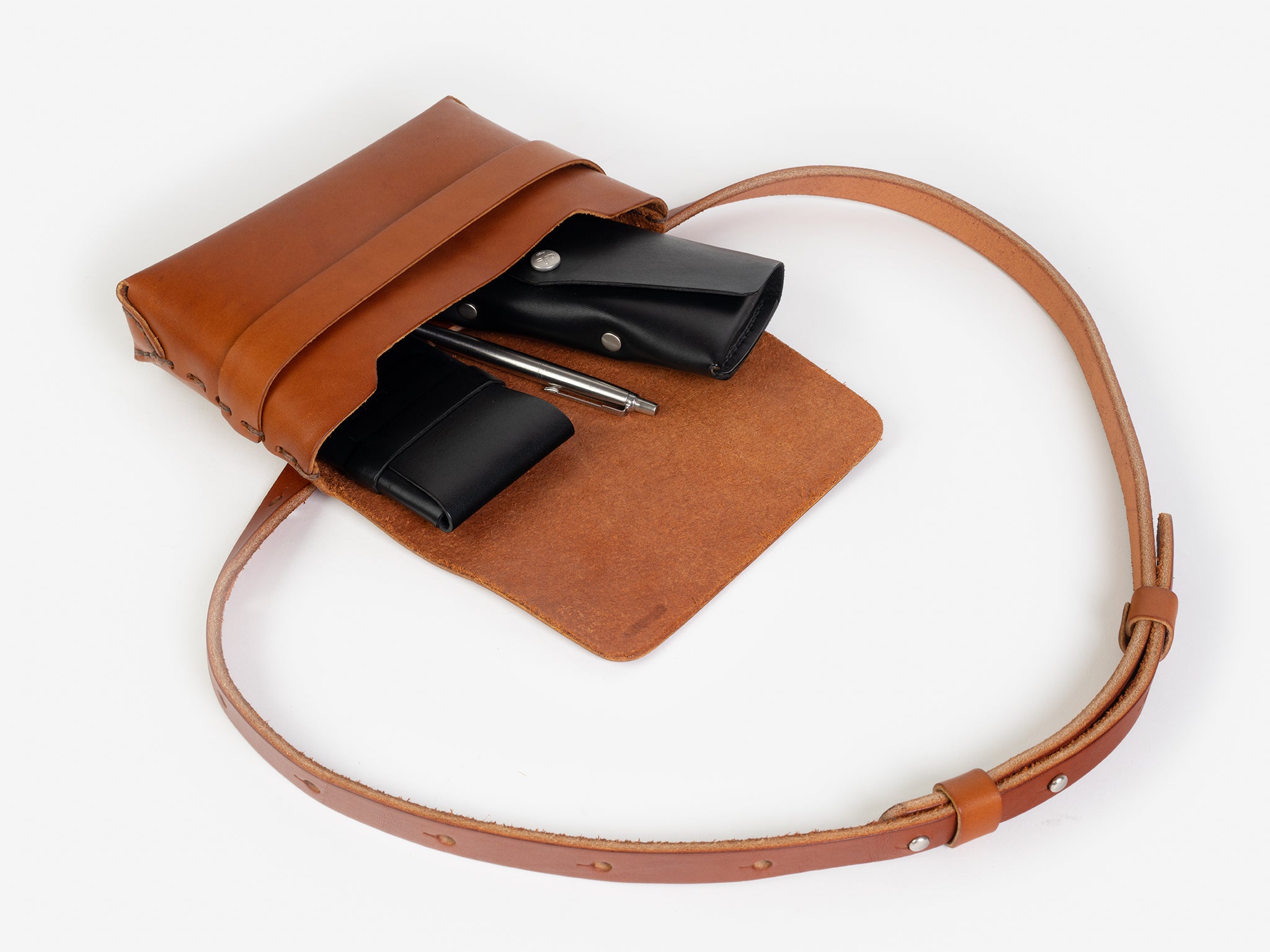 A tan leather crossbody bag with a flap open, revealing a black card case, a silver pen, and a black leather sunglasses case inside. The bag has a long adjustable strap and minimalist design, displayed on a white background.