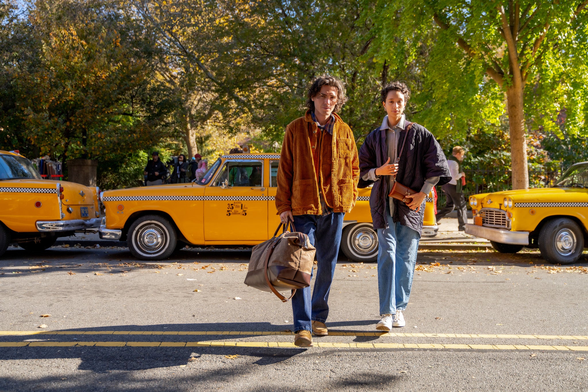 Two young people walk side by side on a sunny street, with one carrying a duffel bag. Behind them are classic yellow taxis and trees with autumn foliage.