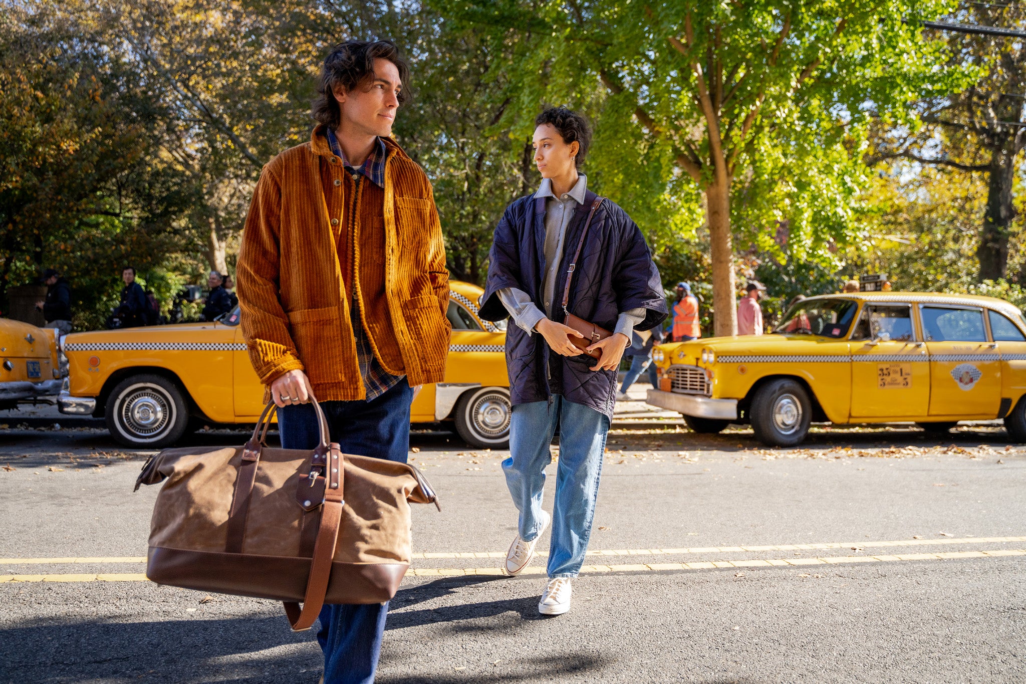 Two people walk on a sunny street lined with classic yellow taxis. One, holding a brown duffel bag, wears a brown corduroy jacket; the other, in a blue coat, carries a book. Trees with green leaves are in the background.
