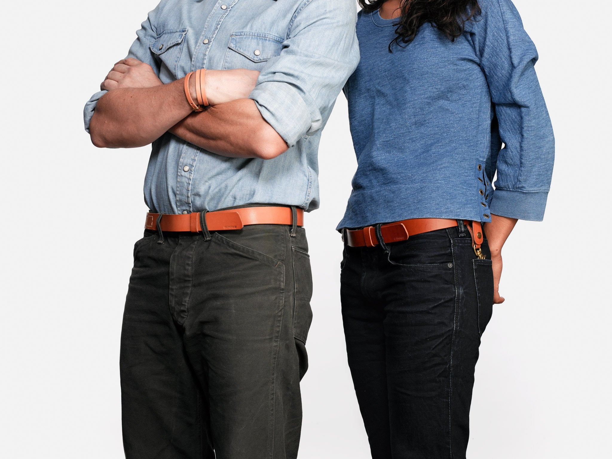 Two people standing side by side with arms crossed and hands behind back, wearing blue tops, dark pants, and brown belts, facing forward against a plain white background. Their faces are not visible.