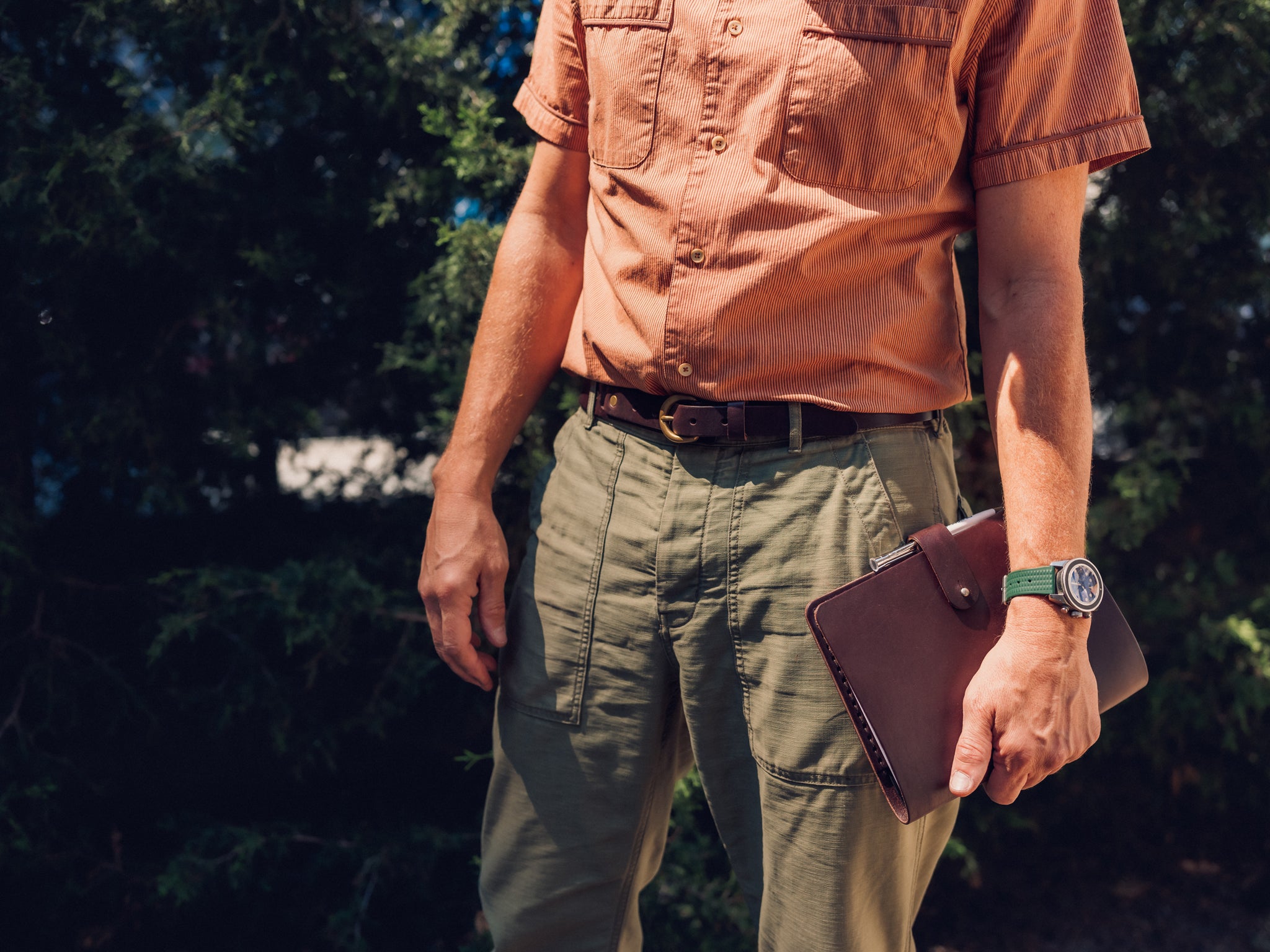 A person wearing a short-sleeve orange shirt and olive green pants stands outdoors, holding a brown leather portfolio in one hand. Trees and greenery are visible in the background.
