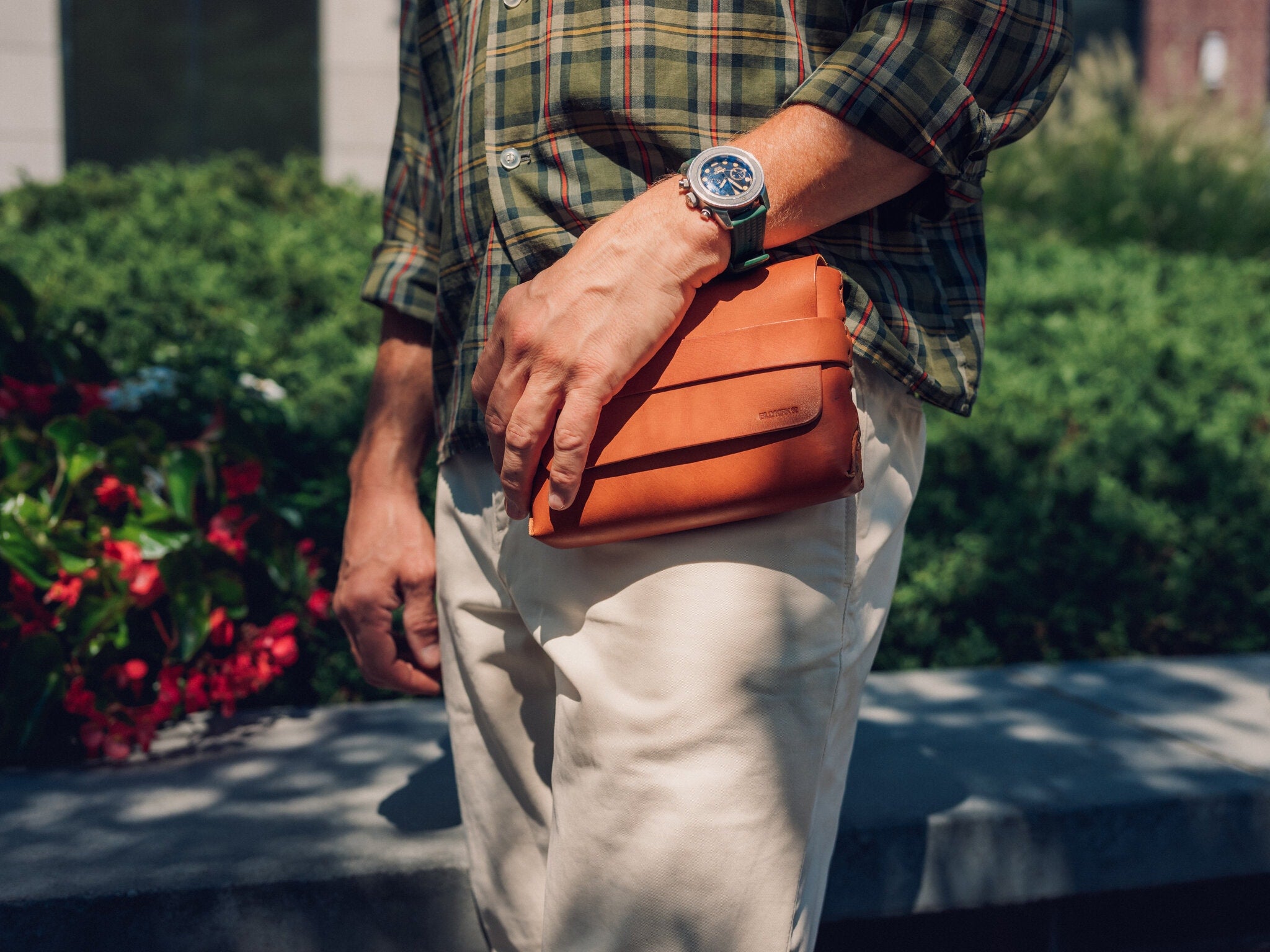 A person wearing a green plaid shirt and beige pants holds a small tan leather bag. They are outdoors near greenery and flowers, and wearing a silver wristwatch.