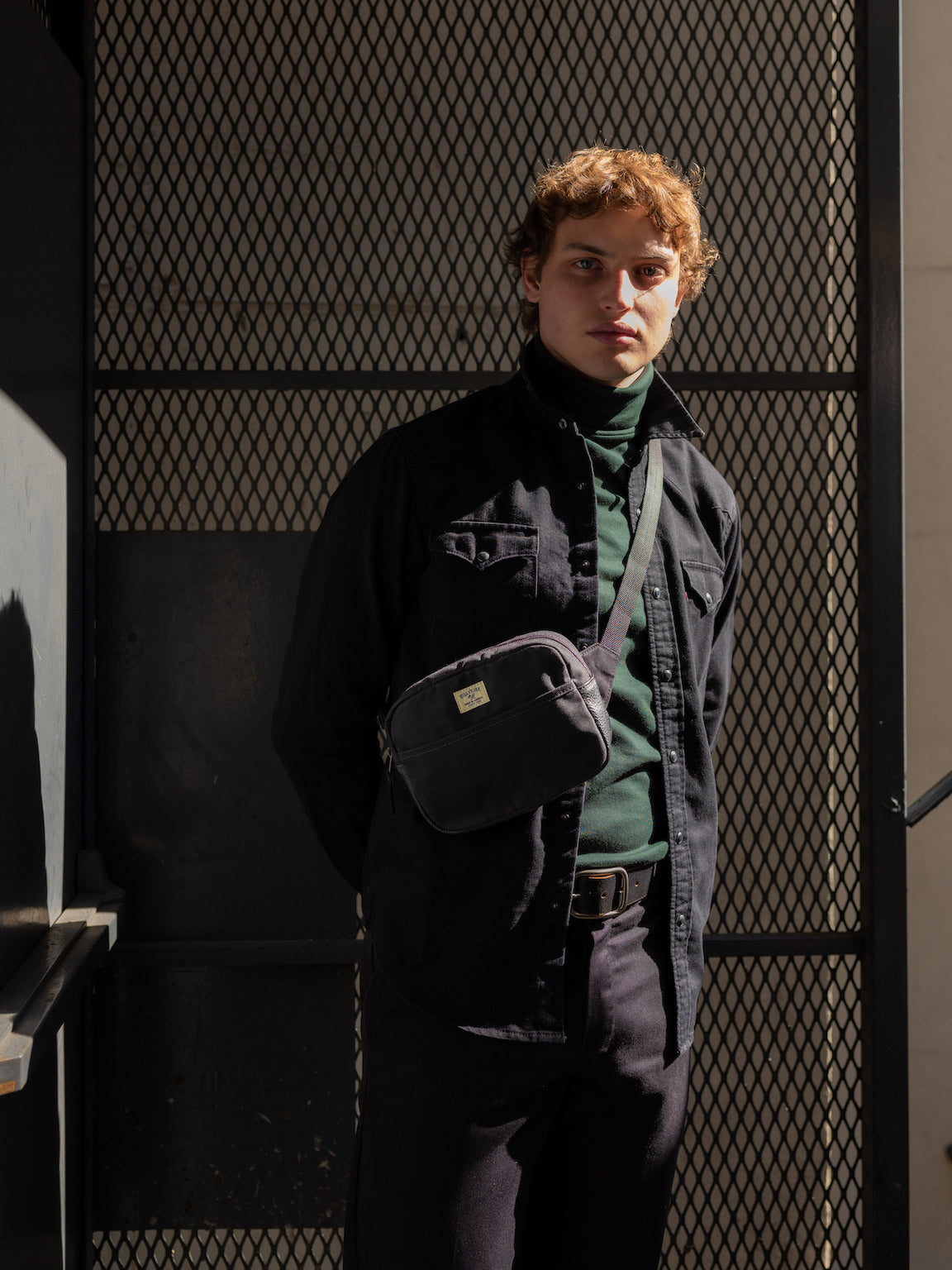 A person with curly hair stands in sunlight against a metal mesh fence, wearing a dark jacket over a green turtleneck, with a black waxed canvas crossbody bag and hands behind their back.