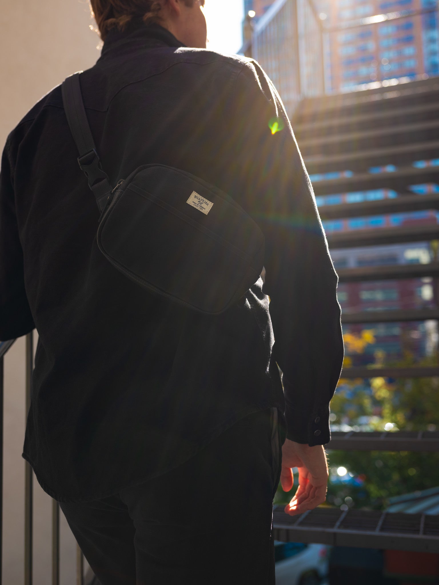A person wearing a black shirt and black waxed canvas crossbody bag stands outdoors near stairs, with sunlight streaming in and city buildings visible in the background.