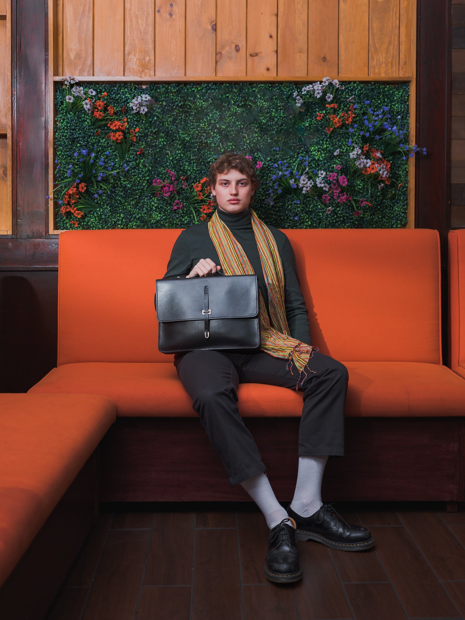 A person sits on an orange couch, holding a black briefcase on their lap. They wear dark clothes, white socks, black shoes, and a striped scarf. Behind them is a green wall with colorful flowers and wooden paneling.