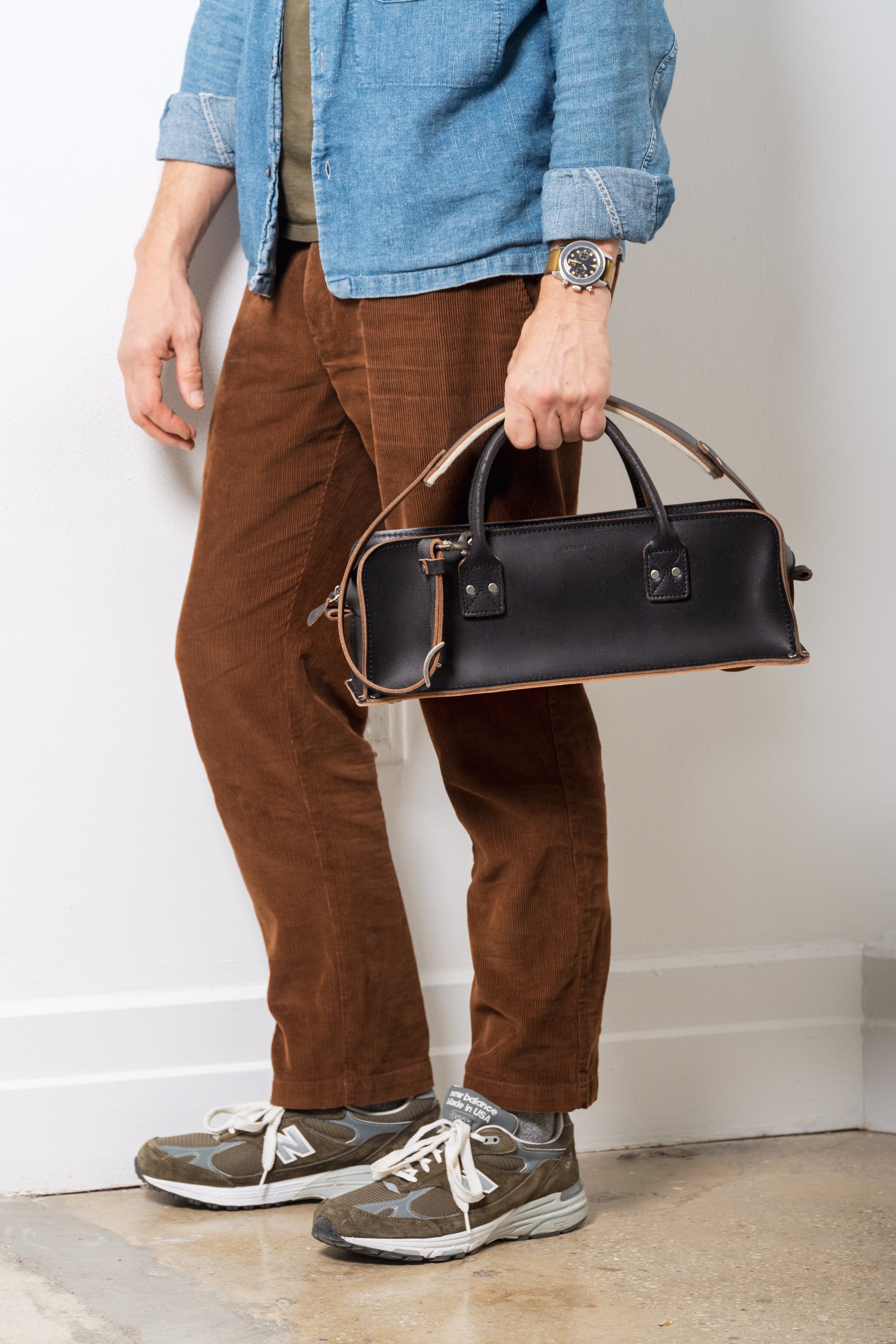 A man in brown pants and blue shirt is holding a black leather tool bag with shoulder strap against a white wall