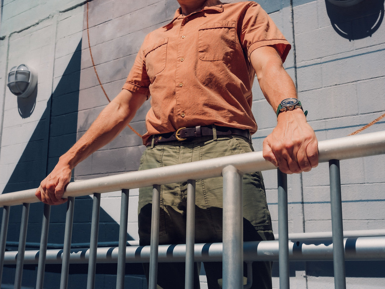 A person in a short-sleeved orange shirt and green pants stands outside, gripping a metal railing with both hands. The photo is taken from a low angle against a painted wall in sunlight.