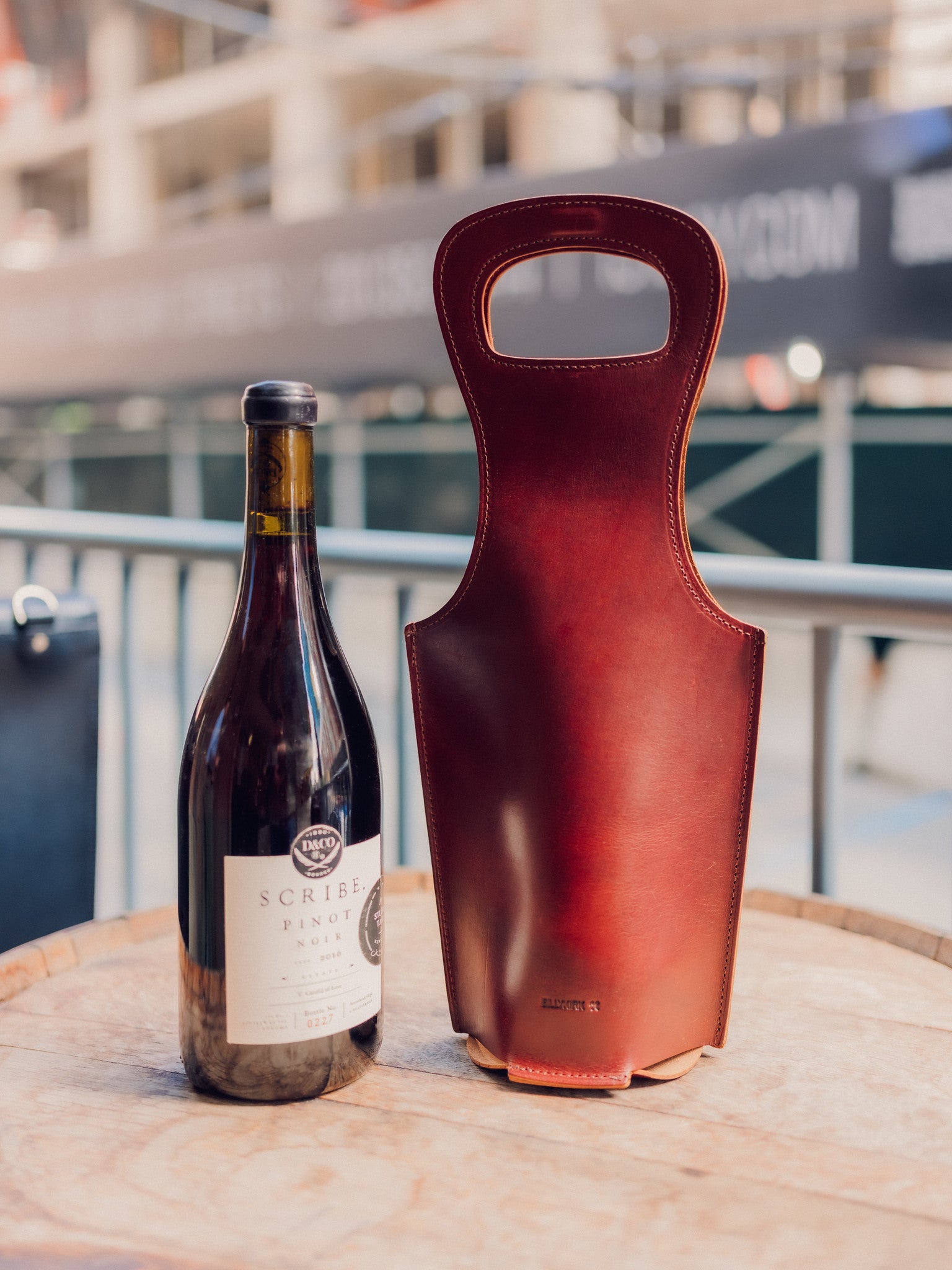 A bottle of Scribe Pinot Noir wine stands next to an elegant brown leather wine carrier on a round wooden table, with a blurred urban background.