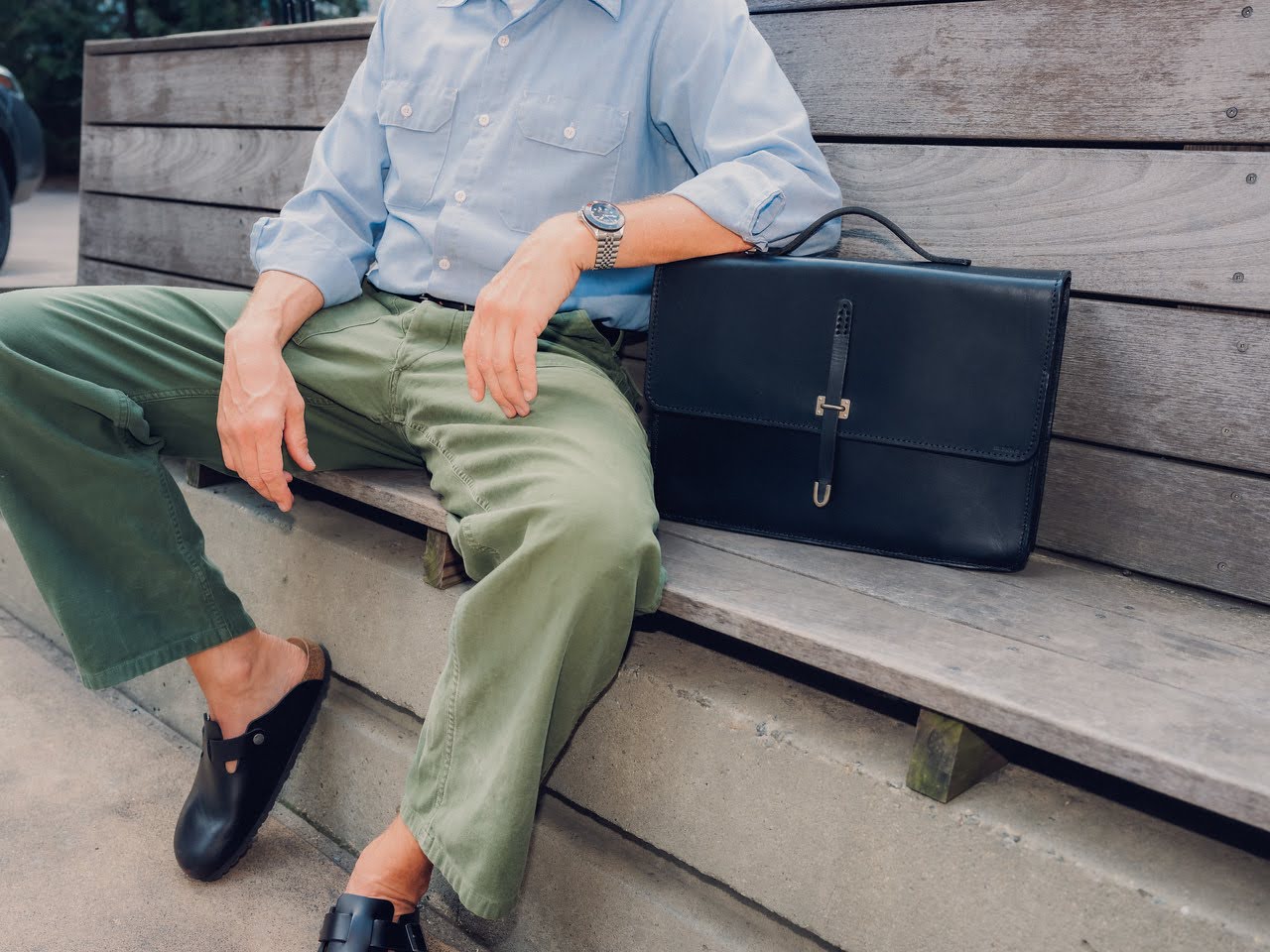 A person wearing a light blue shirt, green pants, and black slip-on shoes sits on a wooden bench with a black leather briefcase beside them. The persons face is not visible.