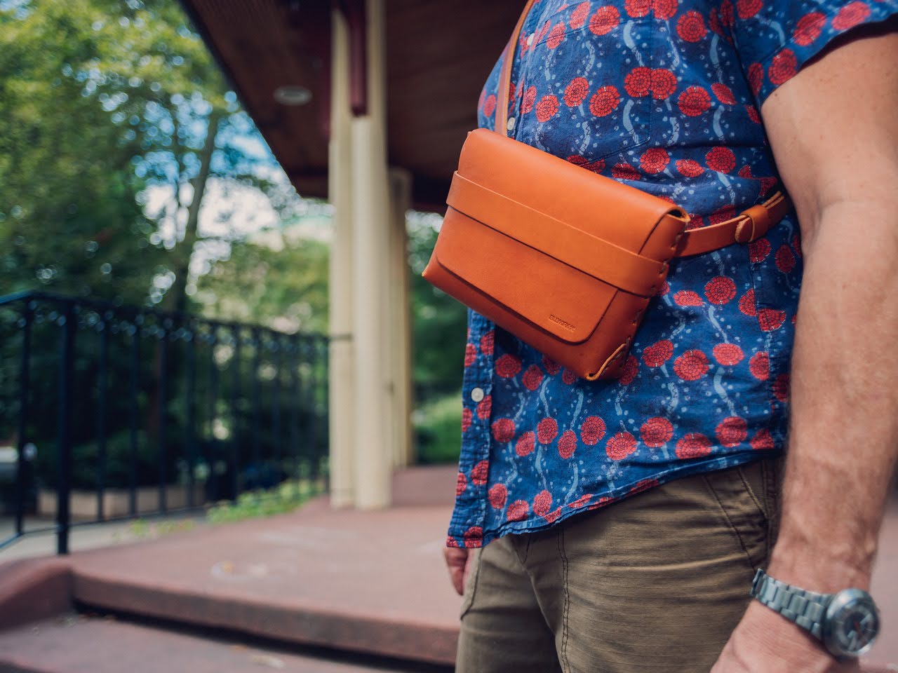 A person wearing a blue shirt with red patterns stands outside, carrying a tan leather crossbody bag and wearing a wristwatch. Trees and a building with columns are visible in the background.