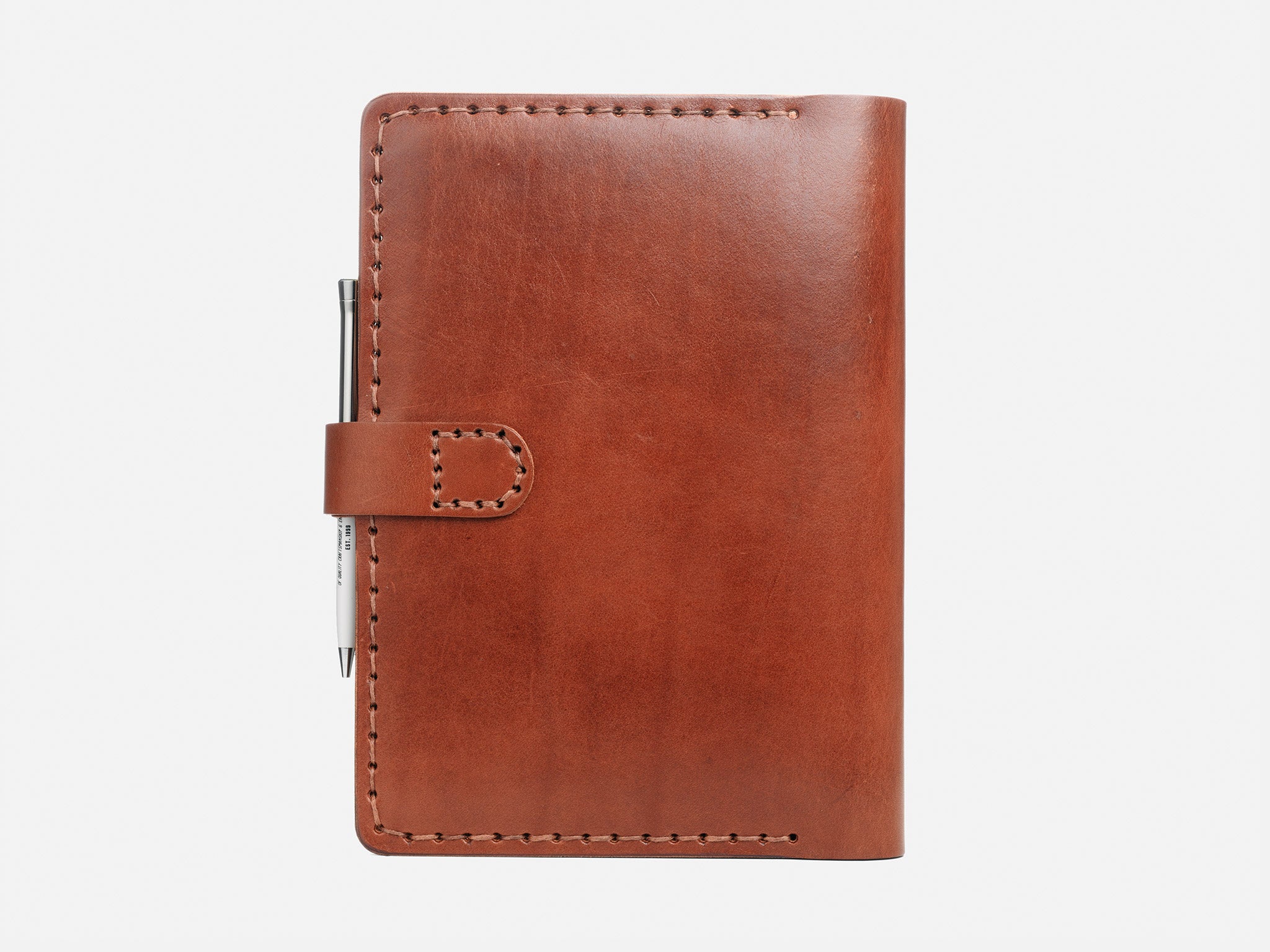 A closed brown leather notebook with visible stitching and a pen tucked into a loop on the left side, placed against a plain white background.