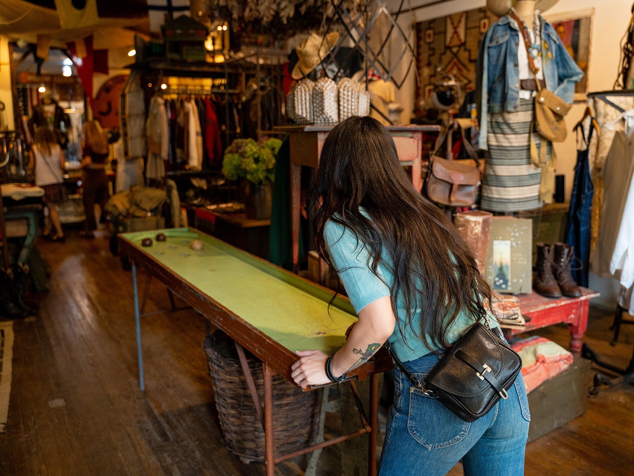 A woman with long dark hair and a black crossbody bag leans over a vintage shuffleboard table in an eclectic, cluttered thrift store filled with clothes, accessories, and decor.