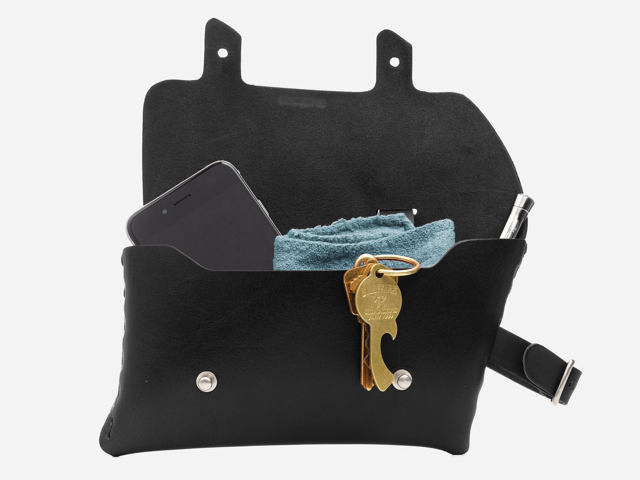 A black leather pouch with its flap open, containing a smartphone, a folded blue cloth, a pen, and a keychain holding several keys and a bottle opener, against a white background.