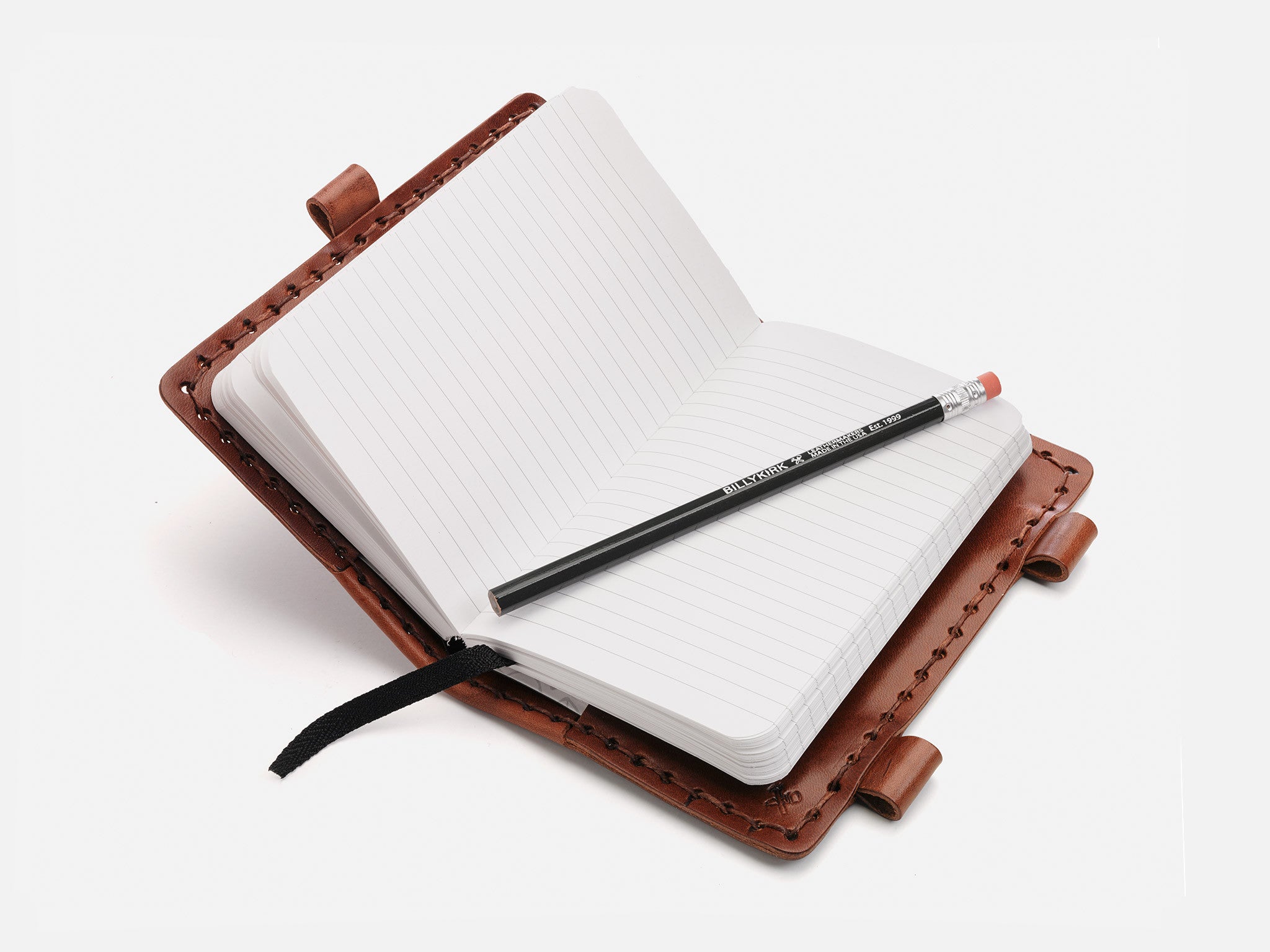 An open, lined notebook with a brown leather cover lies flat, with a black pencil resting diagonally across its pages. A black ribbon bookmark is also visible.
