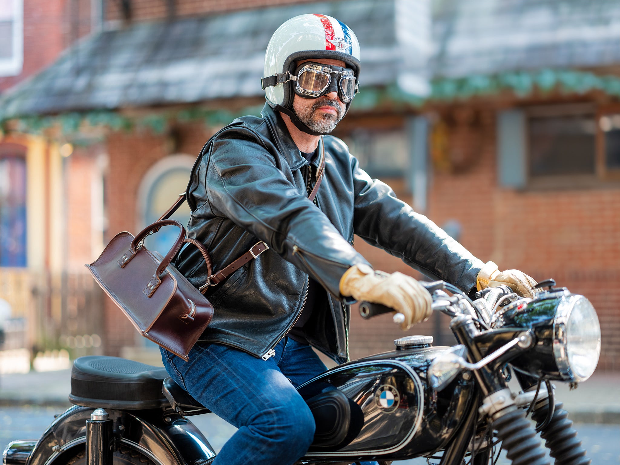 A photo of a man with white helmet, black leather jacket riding a vintage BMW motorcycle with a brown tool bag across his shoulder